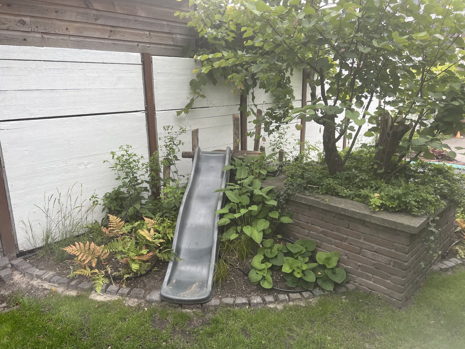 Gemetselde plantenbak met een glijbaan, varens en struiken, met gazon ervoor